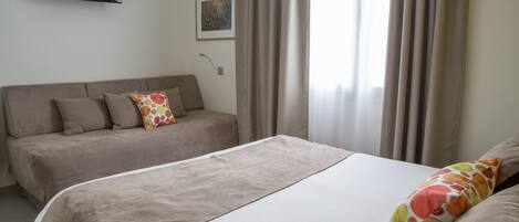 A hotel room with a bed, a sofa, a TV mounted on the wall, and a painting on the wall.