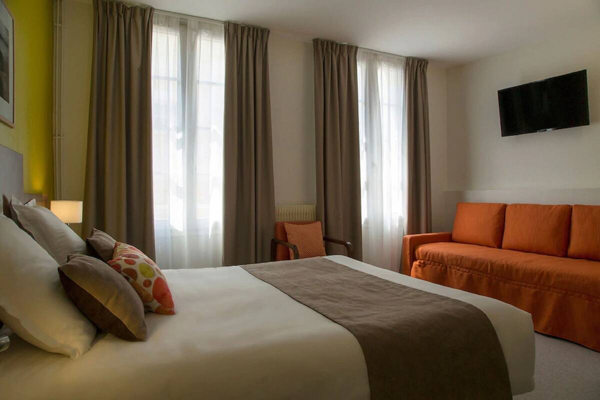A hotel room with a bed, an orange sofa, a TV, and curtains.