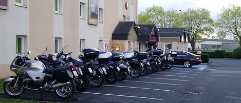 Une rangée de motos garées et une voiture se trouvent sur un parking attenant à un immeuble doté de nombreuses fenêtres et d’une enseigne.