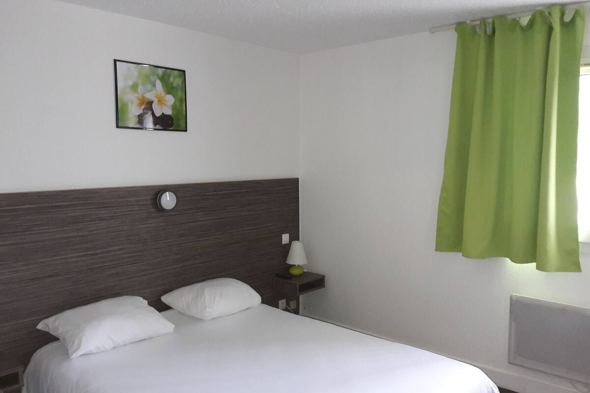 A hotel room with a bed, a nightstand, a lamp, a wall clock, and a window with green curtains.