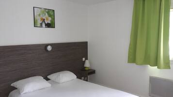 A hotel room with a bed, a nightstand, a lamp, a wall clock, and a window with green curtains.