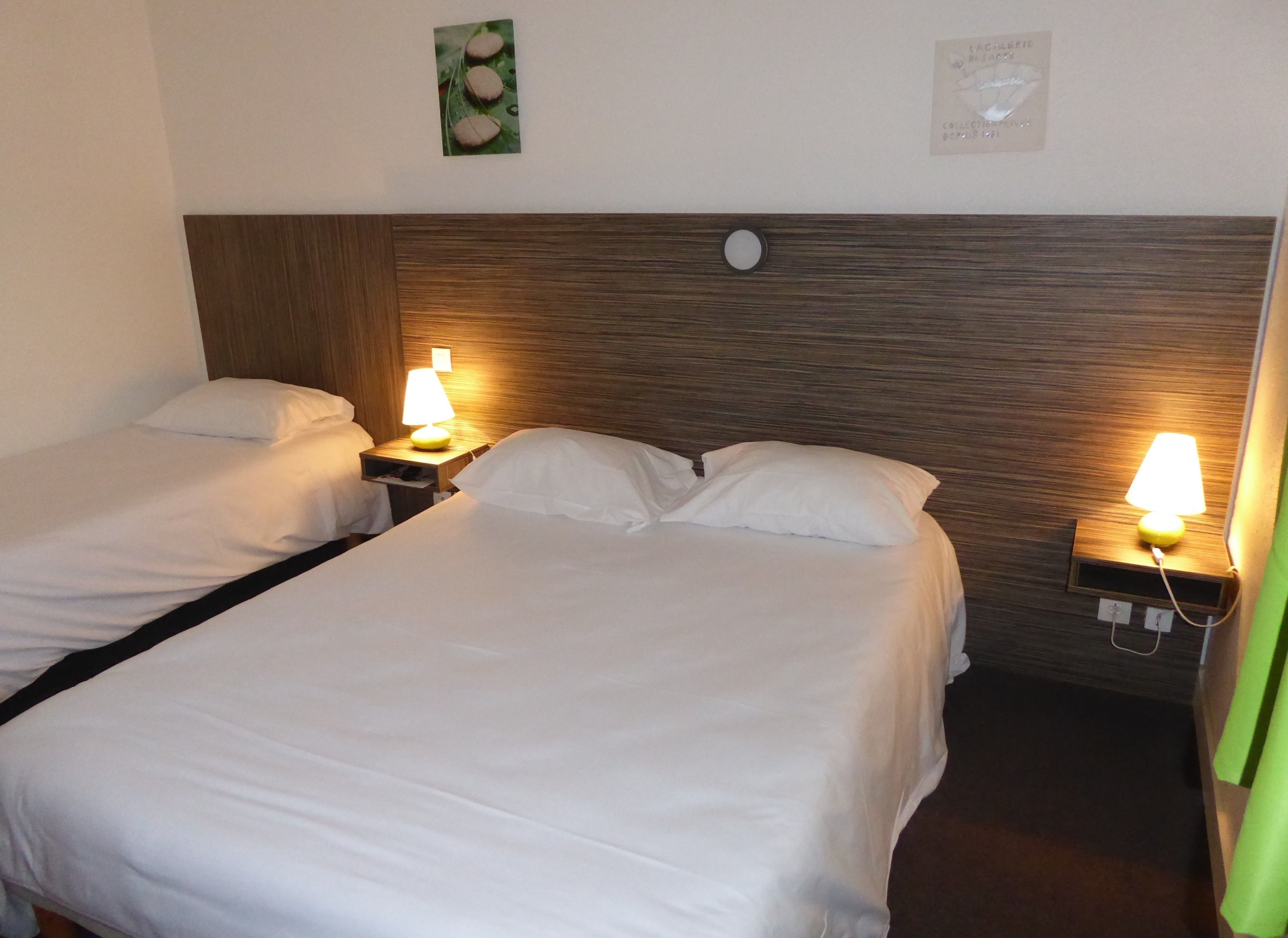 A hotel room with two single beds, each with a white bedspread and matching pillows. There are bedside tables with lamps, a clock on the wall, and a small picture frame.