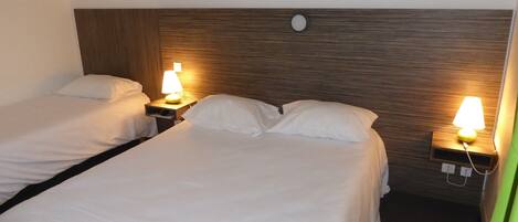 A hotel room with two single beds, each with a white bedspread and matching pillows. There are bedside tables with lamps, a clock on the wall, and a small picture frame.