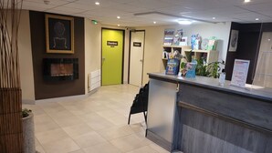 A hotel reception area with a reception desk, a framed artwork on the wall, and doors labeled 'Chambres' and 'Toilets'.