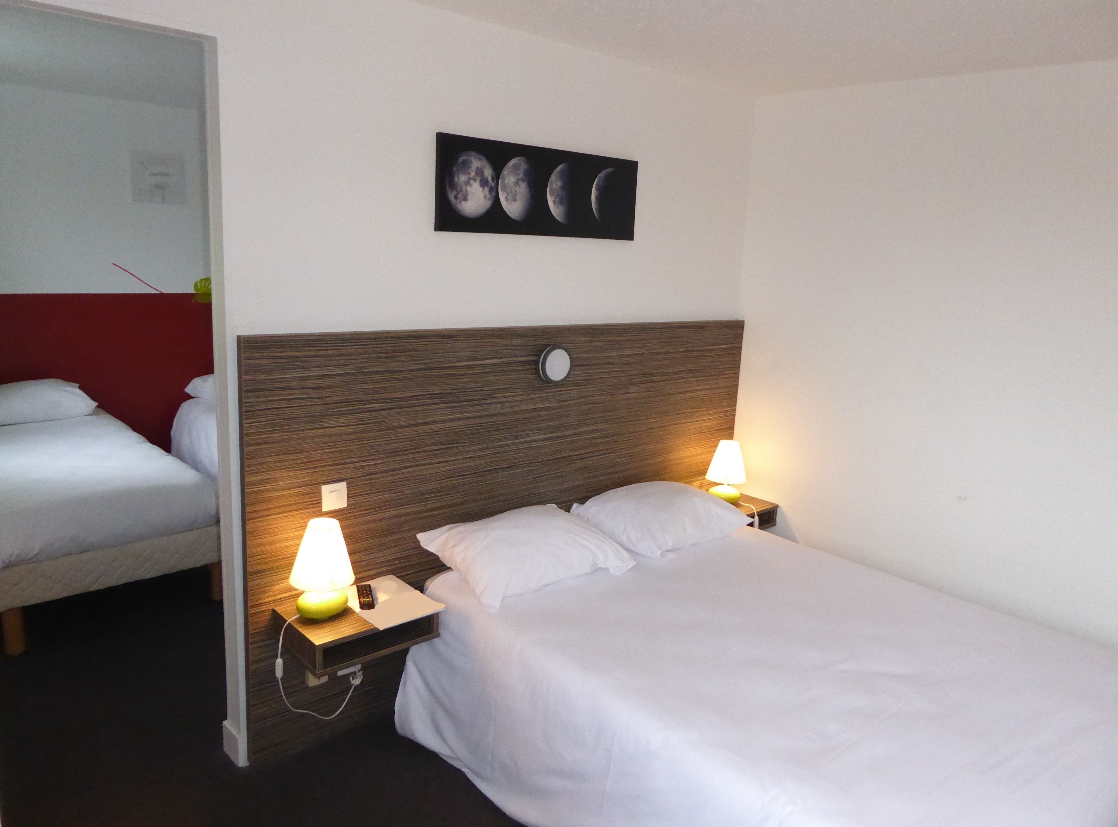A hotel room with two beds, a wooden headboard, bedside lamps, a wall-mounted clock, and a framed picture of the moon phases.