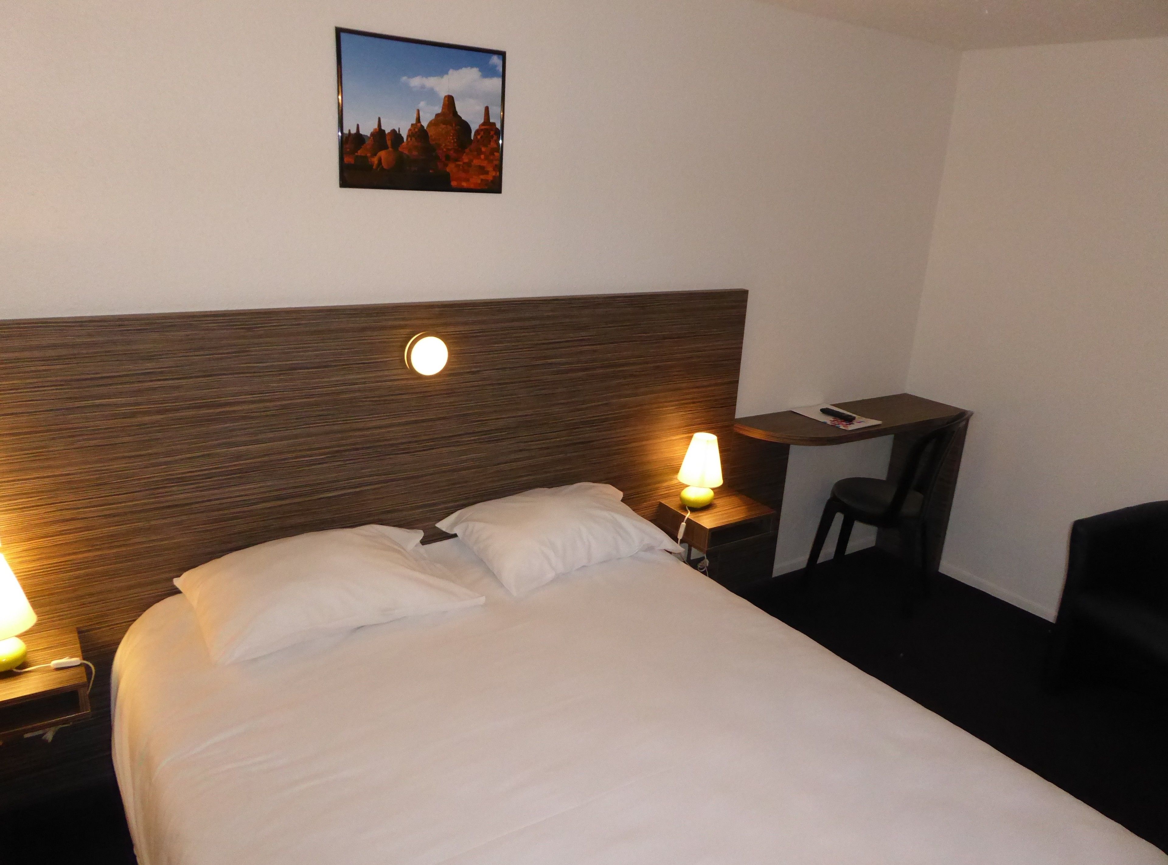 Ein Hotelzimmer mit einem Bett, zwei Nachttischlampen, einem Schreibtisch mit Stuhl und einem gerahmten Bild an der Wand.