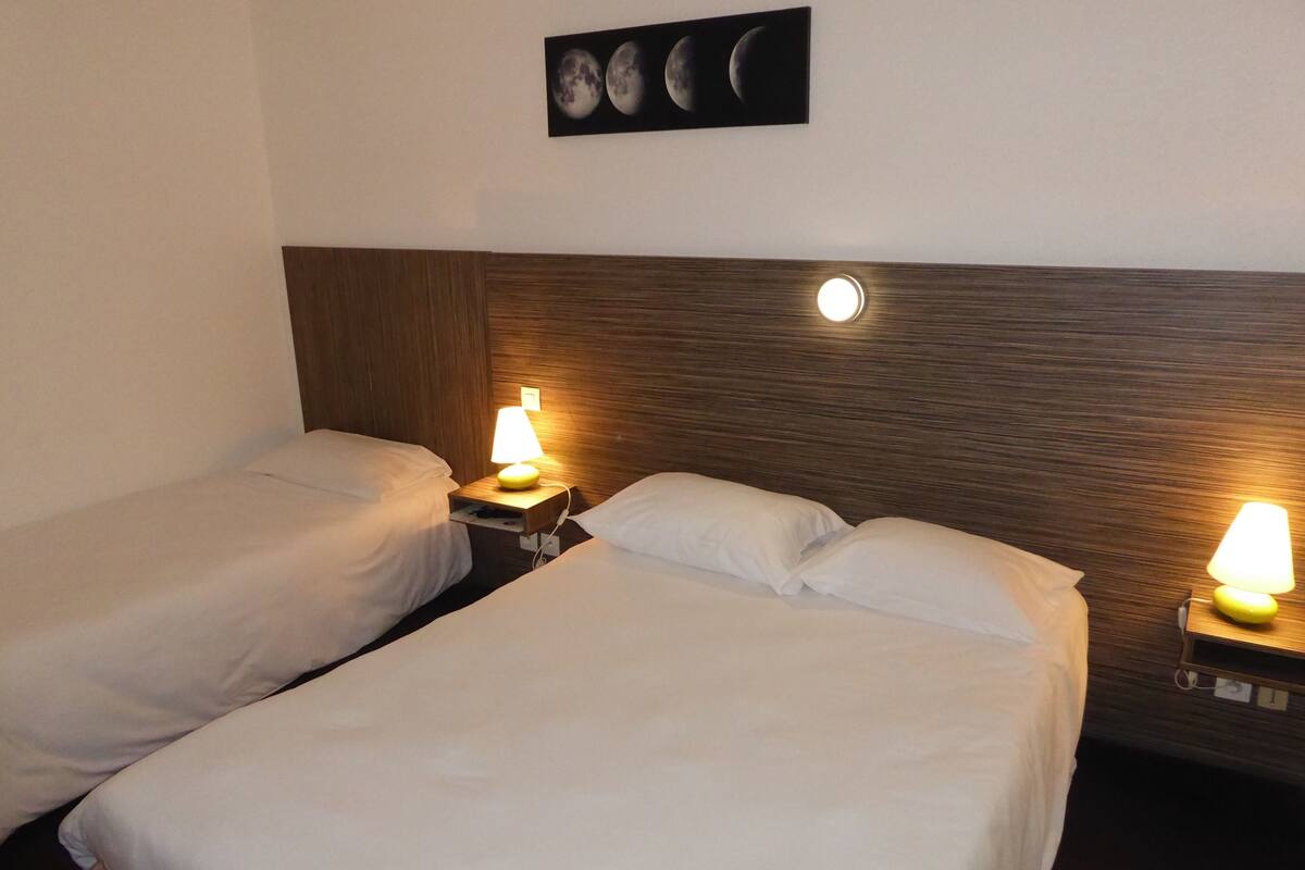 A hotel room with two single beds, each with a bedside table and a lamp. There is a wall-mounted picture above the beds.