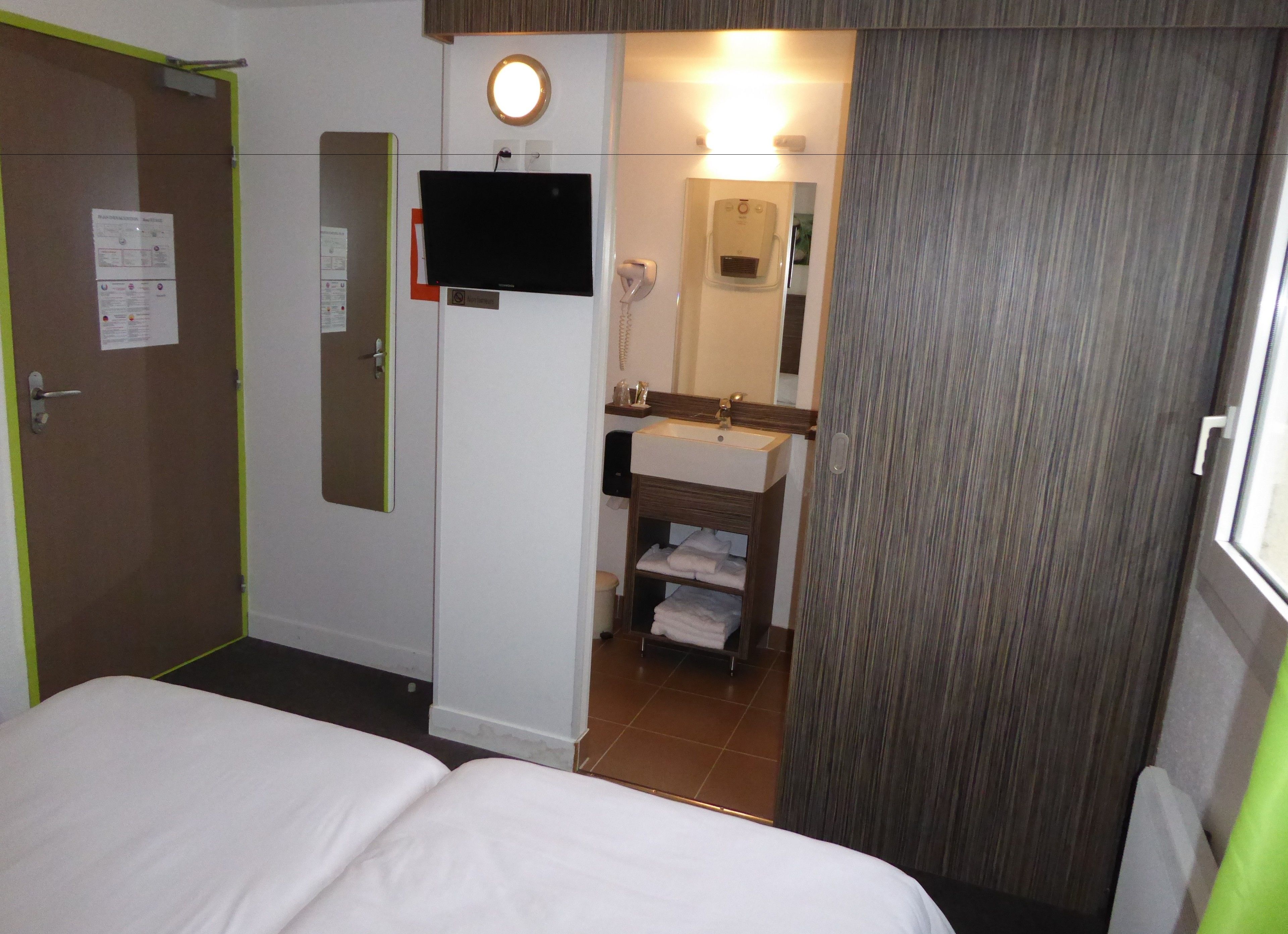 Habitación de hotel con cama, baño con lavamanos y espejo, y una puerta con un tablón de anuncios.