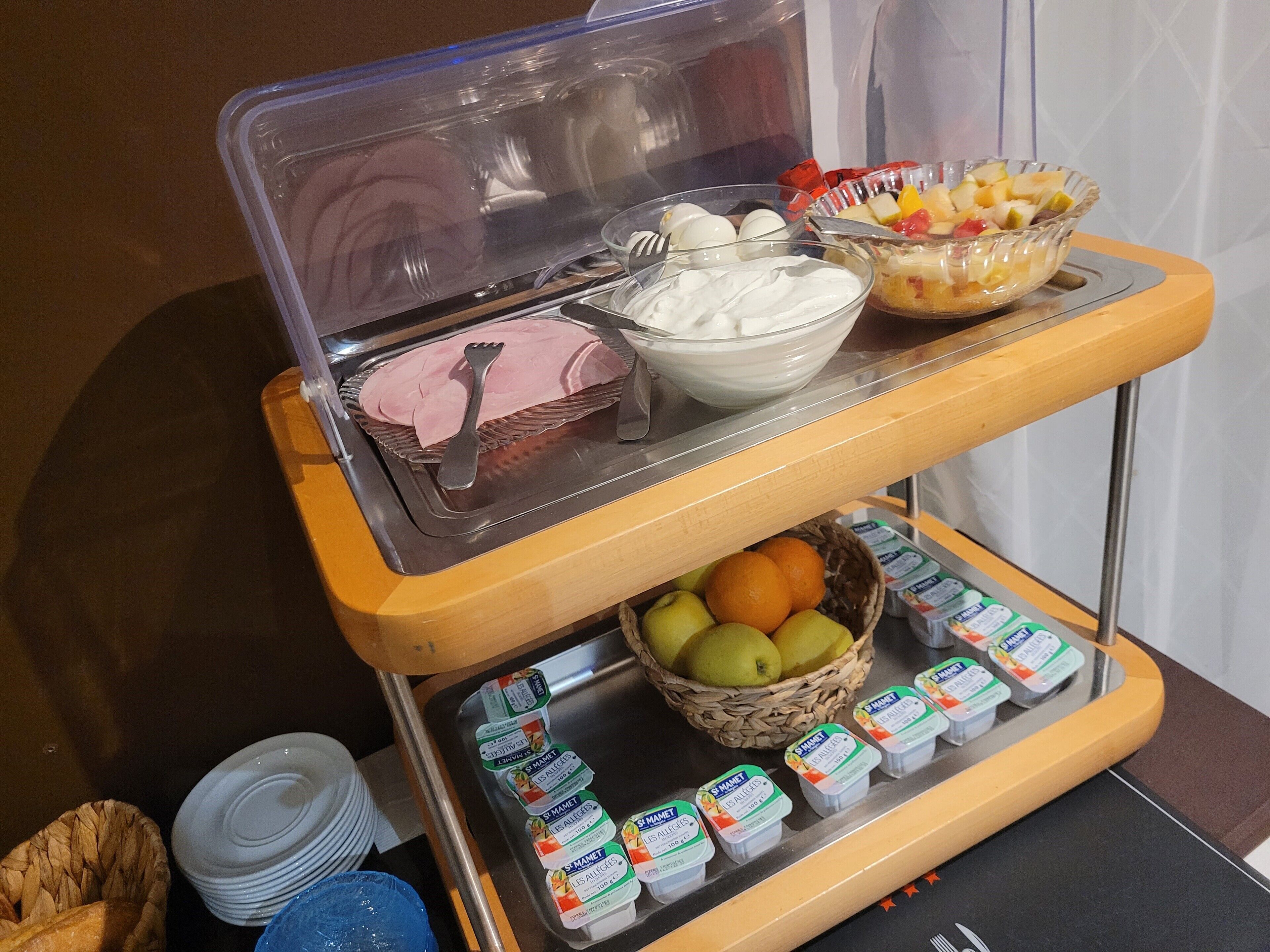 A buffet setup with a variety of food items including a bowl of fruit, a container of yogurt, and a tray of sliced ham.