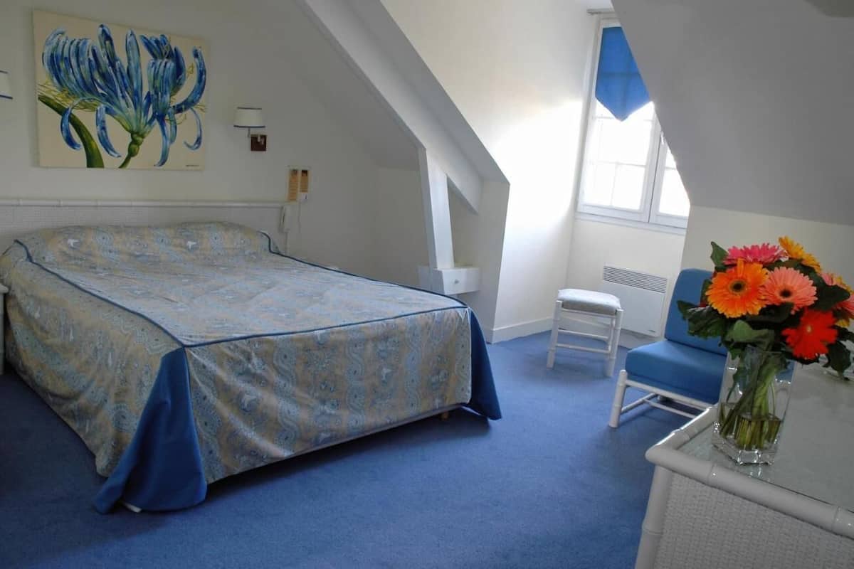 Un dormitorio con cama, una silla, un escritorio, un florero con flores y un cuadro en la pared.