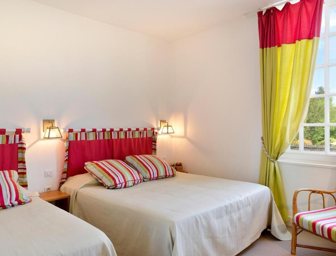 A room with two beds, each with striped pillows, a window with yellow curtains, and wall-mounted lamps.