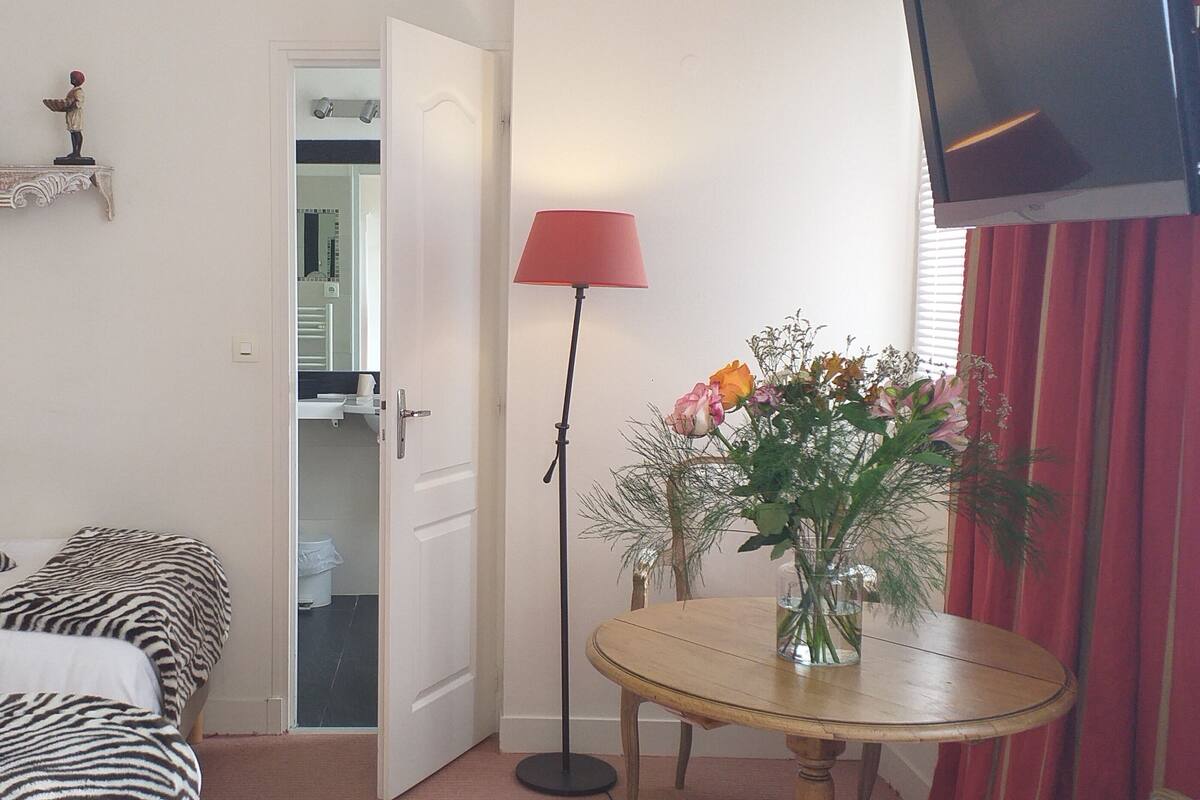 Un dormitorio con dos camas, una mesita con un florero y una lámpara roja, además de un televisor en la pared.