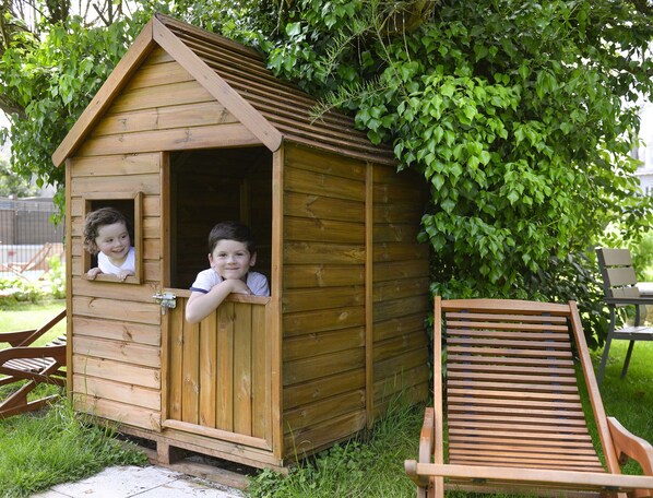 Une maisonnette en bois avec un toit à deux versants et une porte, entourée de verdure.