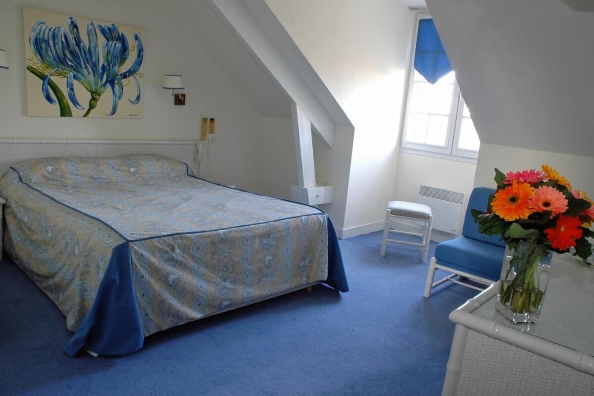 A bedroom with a bed, a chair, a desk, a vase with flowers, and a painting on the wall.