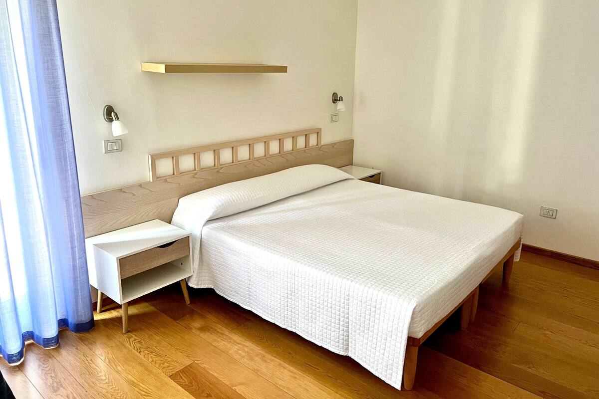 A neatly made bed in a hotel room with a wooden headboard, a bedside table, and a wall-mounted shelf.