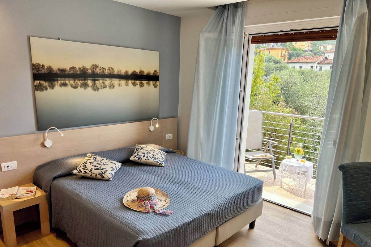 A modern hotel room with a large bed, a painting of a lake, a balcony with a table and chairs, and a view of the surrounding area.