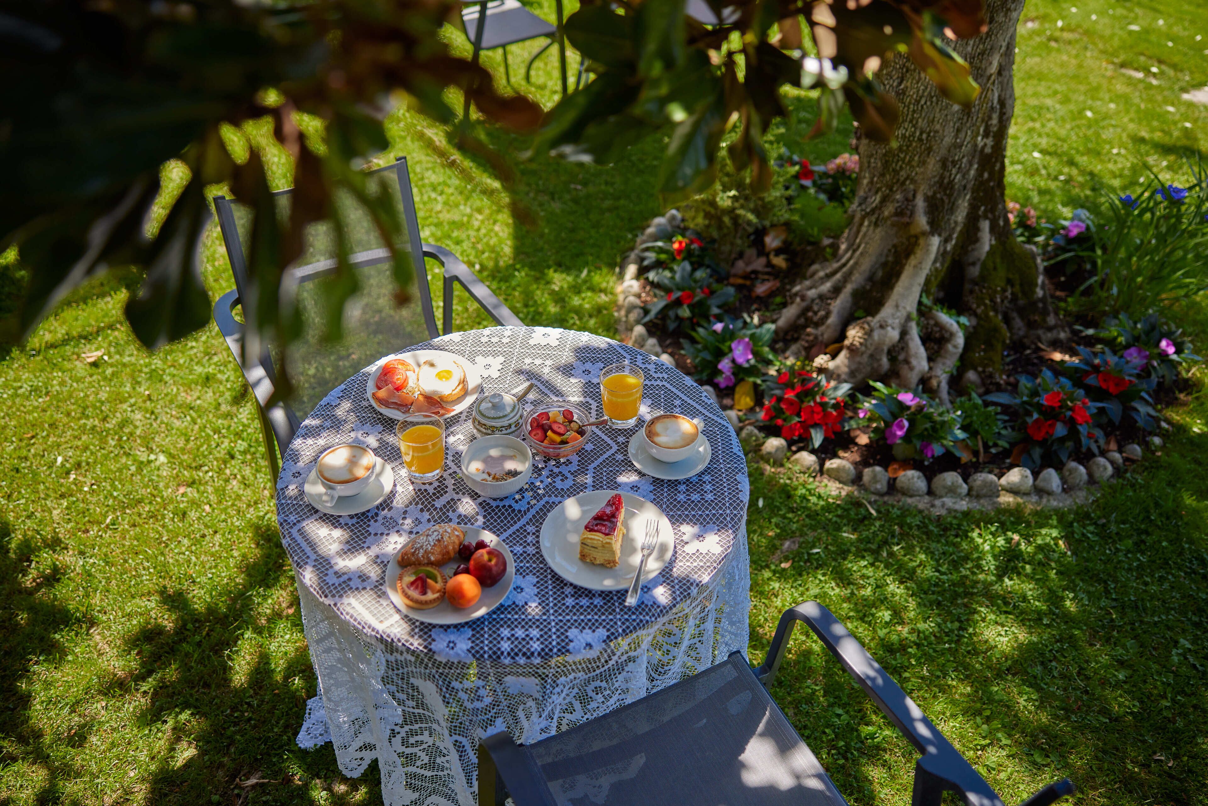 A round table with a lace tablecloth, surrounded by chairs, set outdoors with a garden and a tree in the background.
