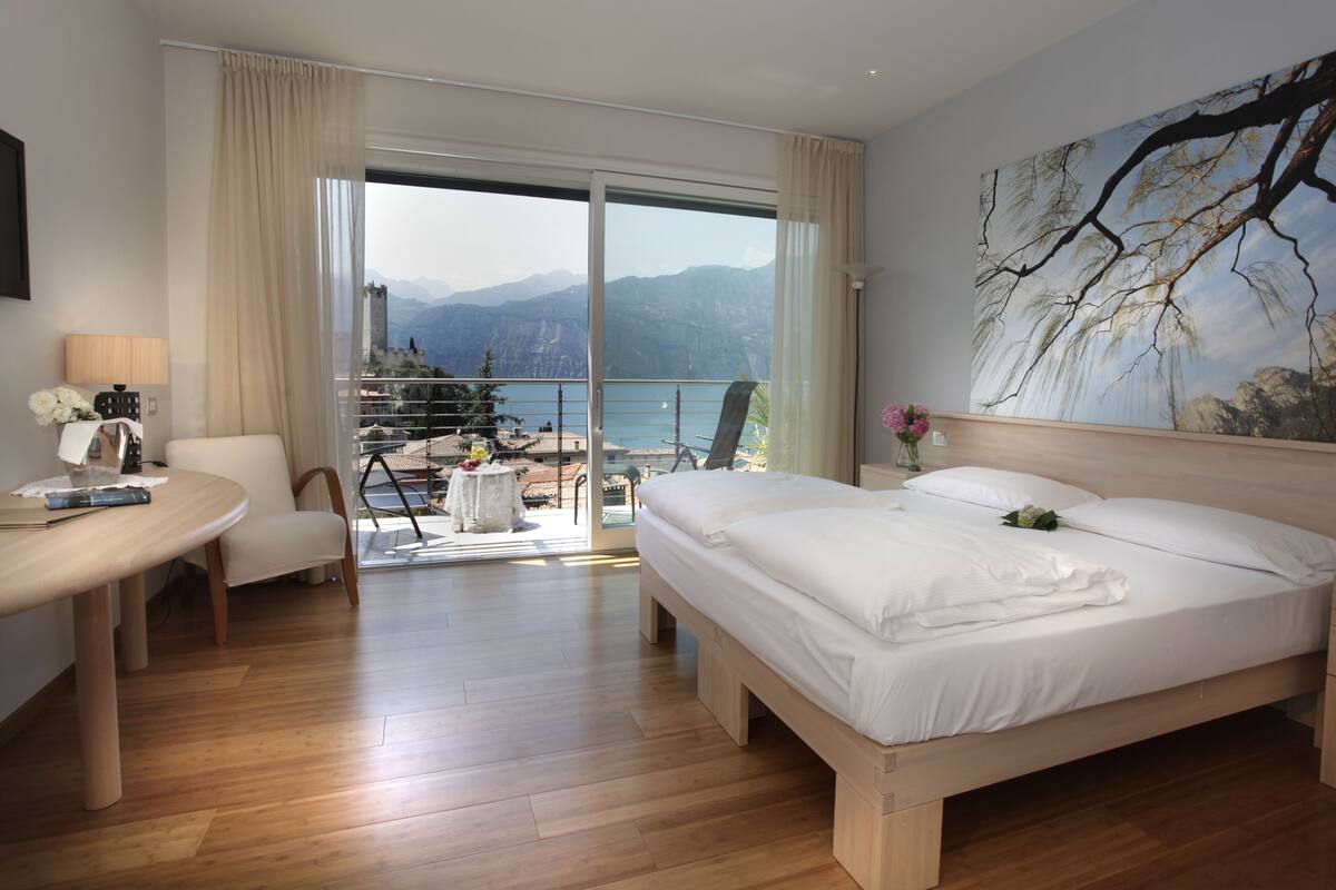 A hotel room with a large bed, a desk, a chair, and a view of mountains through a balcony.