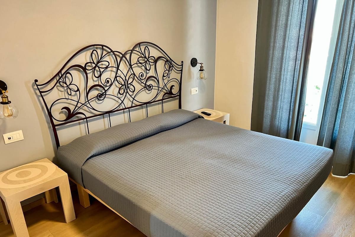 A neatly made bed with a grey quilt, a metal headboard with floral patterns, wooden nightstands, and a bedside lamp.