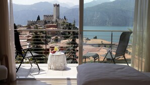 A balcony with a bed, two chairs, and a table with fruit. A castle and mountains are visible in the background. - Hotel Ariston (Malcesine)