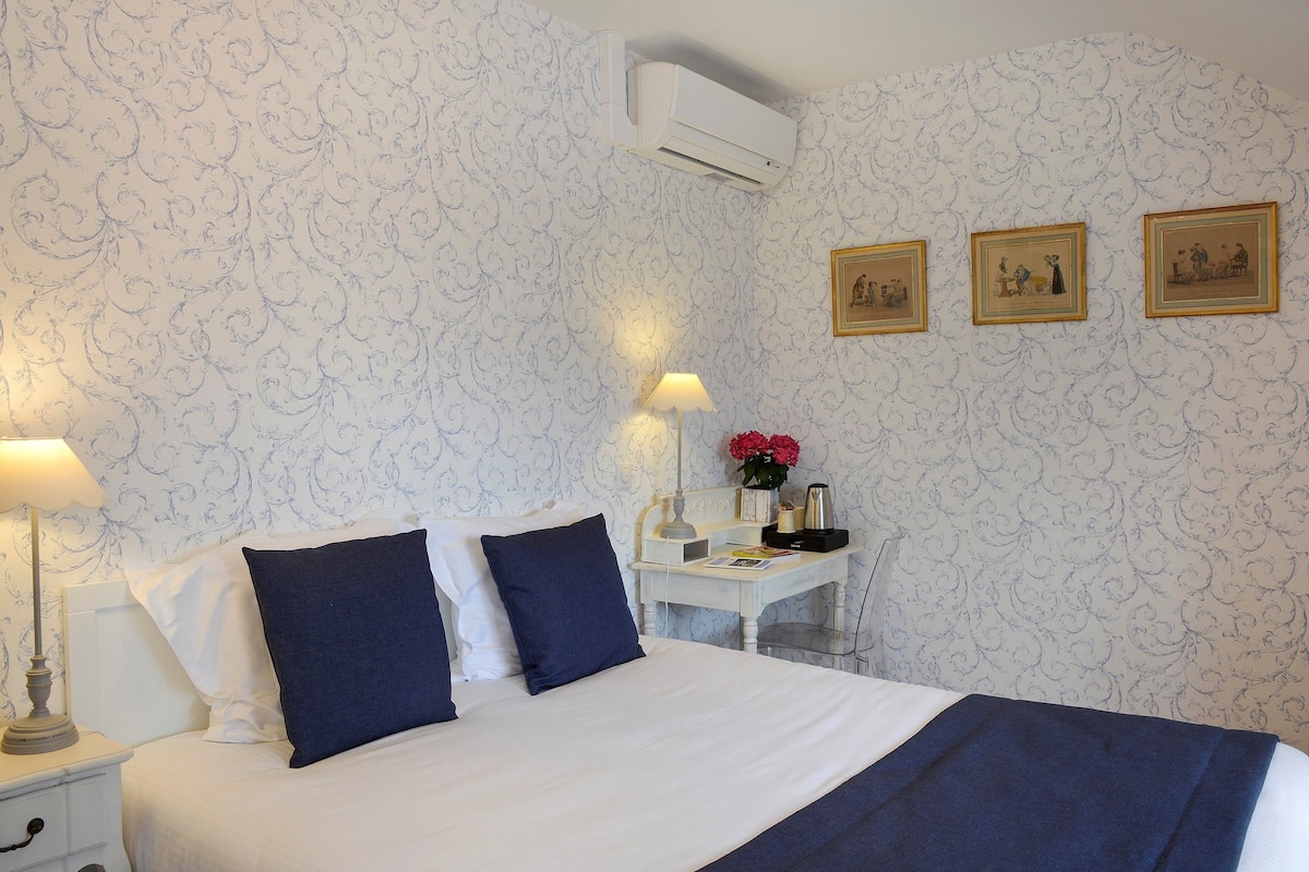Una cama bien hecha con una combinación de colores blanco y azul oscuro, una mesita de noche con una lámpara, y un escritorio con un florero de flores.