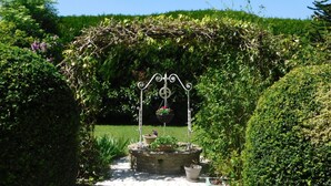 Un sendero de jardín que conduce a una fuente de piedra con un arco de metal y cestas de flores colgantes.