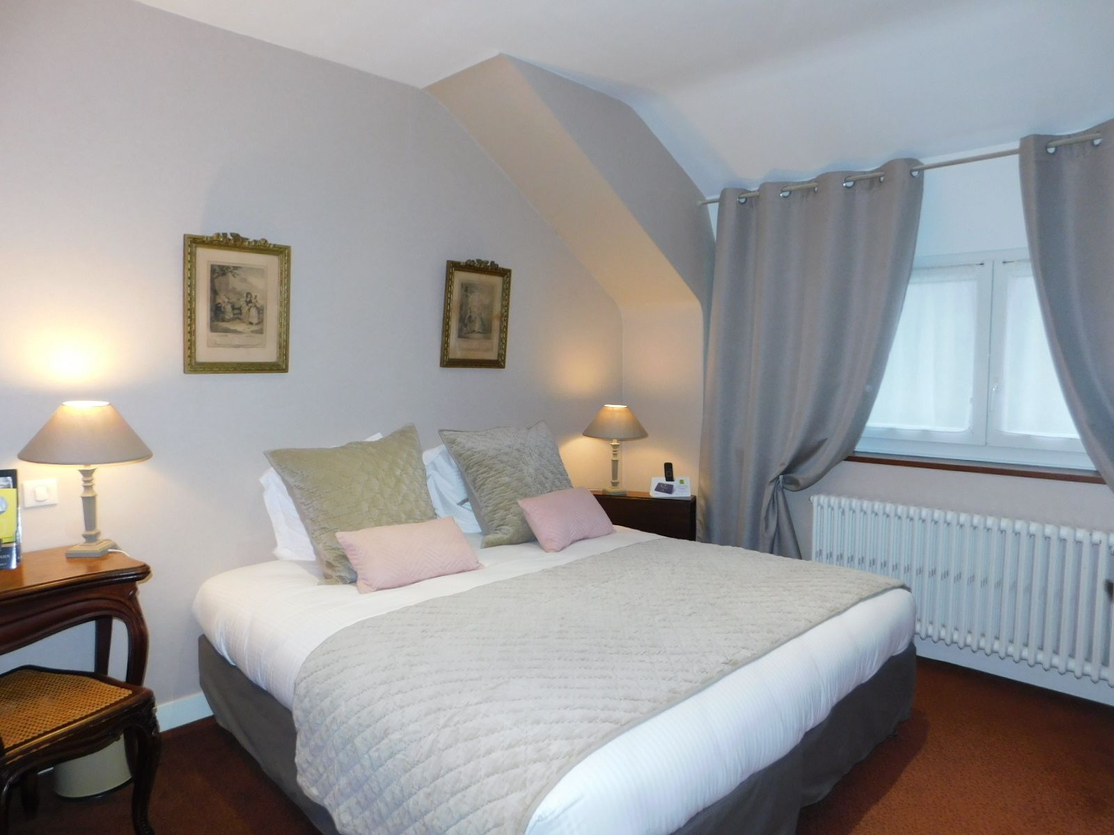 A bedroom with a large bed, two bedside tables with lamps, a radiator, and two framed pictures on the wall.