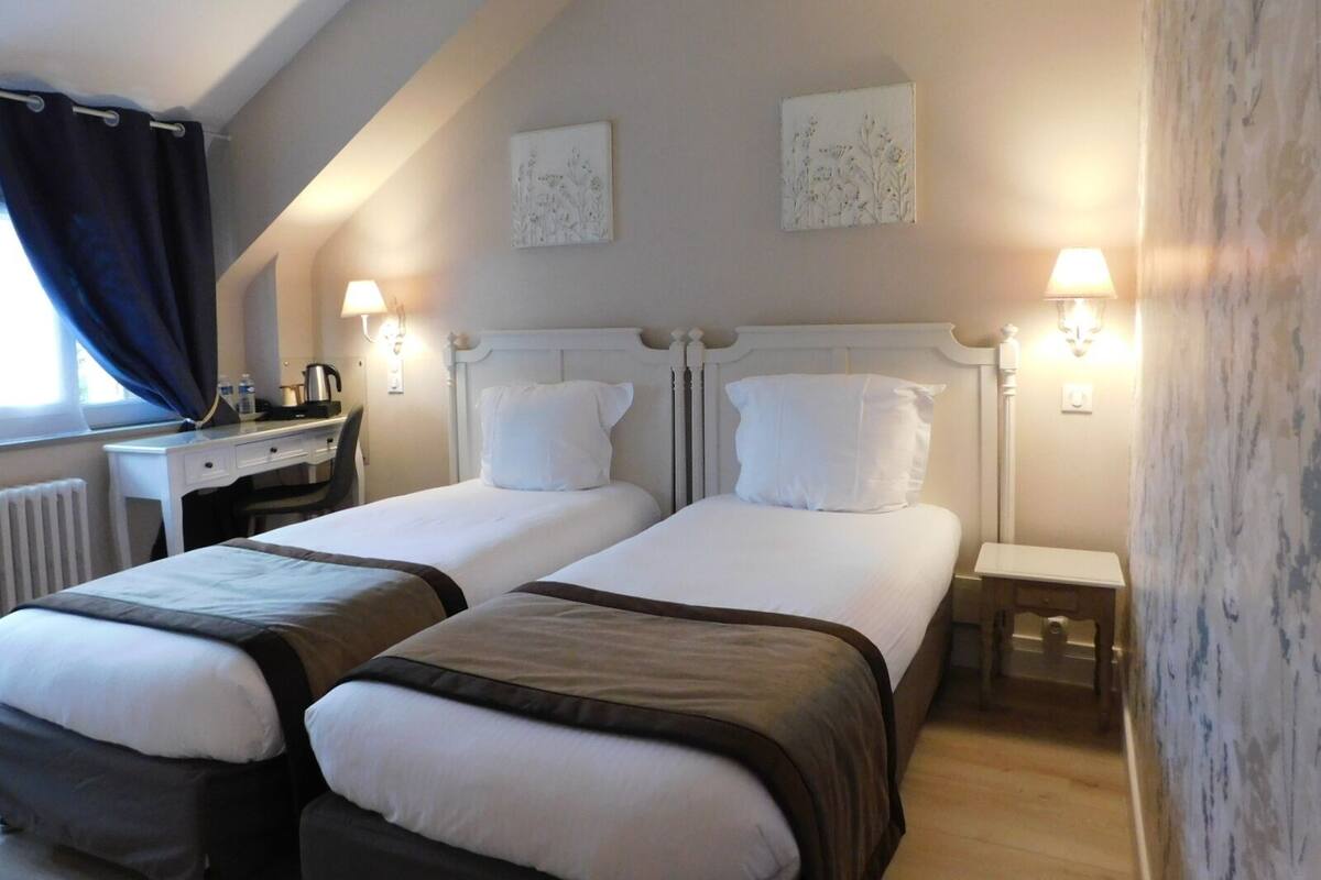 Habitación de hotel con dos camas, un escritorio y una silla. Hay lámparas fijadas a la pared y un radiador.