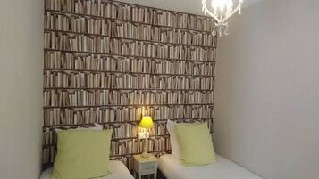 A room with two beds, a wall of books, a chandelier, and a nightstand with a lamp.
