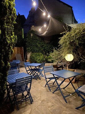 Un espace extérieur aménagé en terrasse, avec des tables et des chaises en métal bleu, des guirlandes lumineuses et une lanterne.