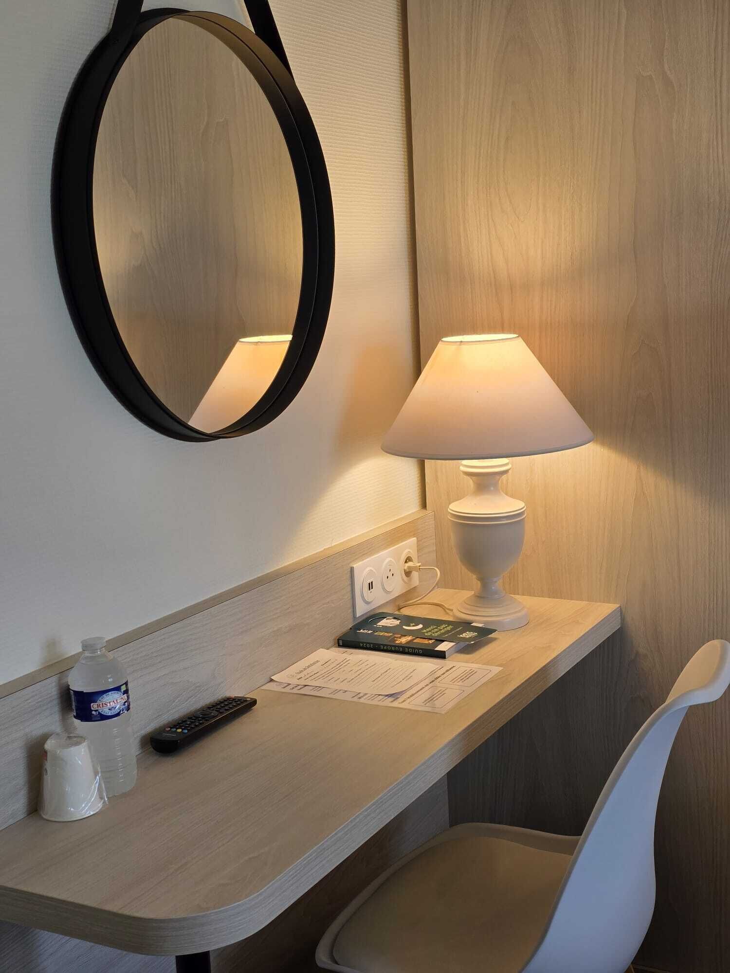 A hotel room with a wooden desk, a white chair, a round mirror, a table lamp, a remote control, a water bottle, and a book.