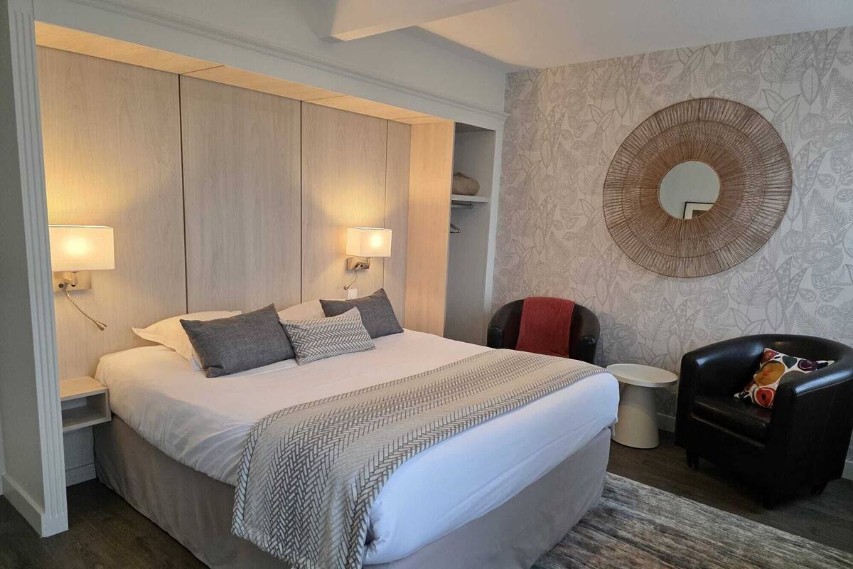 A modern hotel room with a large bed, two bedside lamps, a round mirror, a small round table, and a leather armchair.