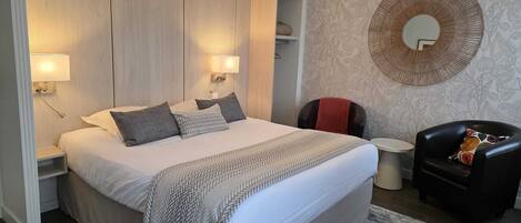 A modern hotel room with a large bed, two bedside lamps, a round mirror, a small round table, and a leather armchair.