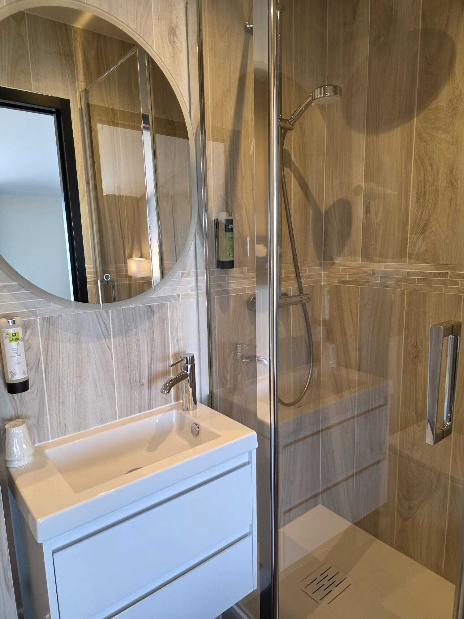 Un baño moderno con ducha envuelta en vidrio, tocador blanco con espejo y pared de madera.