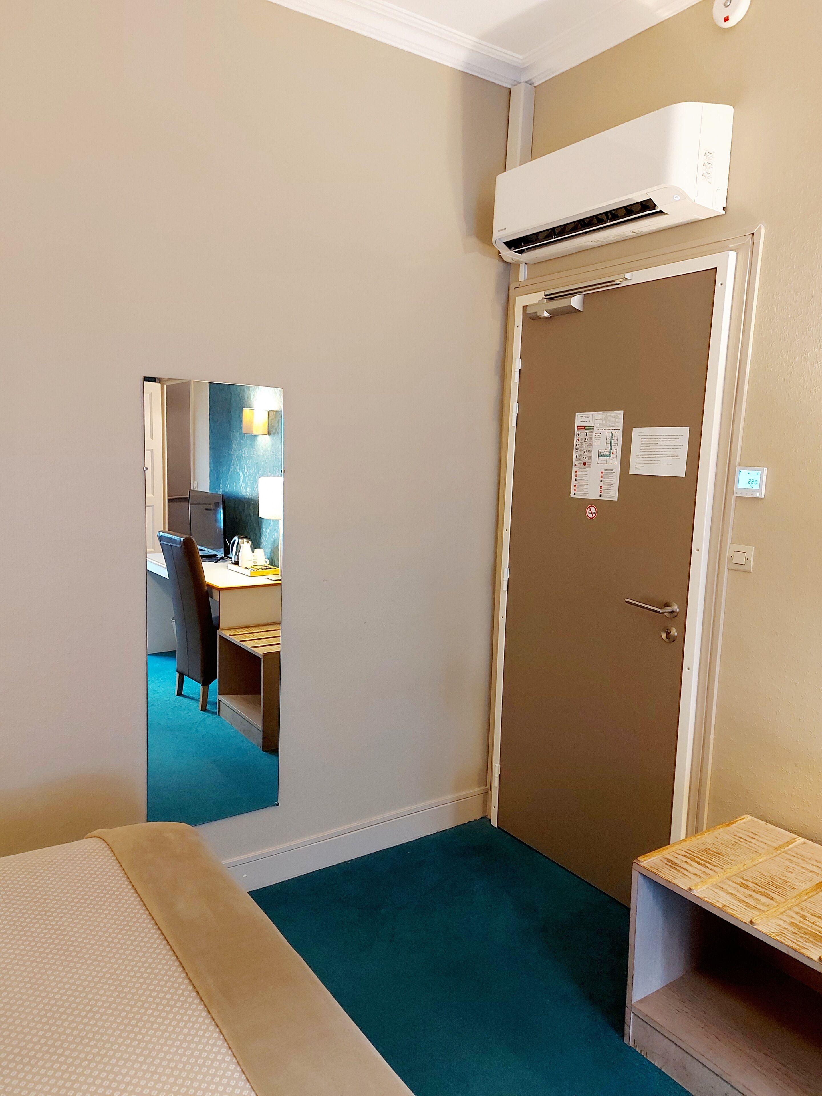 Habitación de hotel con cama, escritorio con silla, equipo de aire acondicionado, puerta con un letrero y una pequeña mesa de madera.