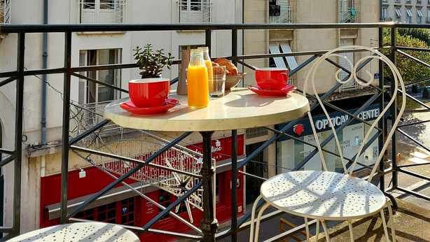 A balcony with a table set for breakfast, including orange juice and croissants, overlooking a building with multiple windows and balconies. - Hotel Mondial (Tours)