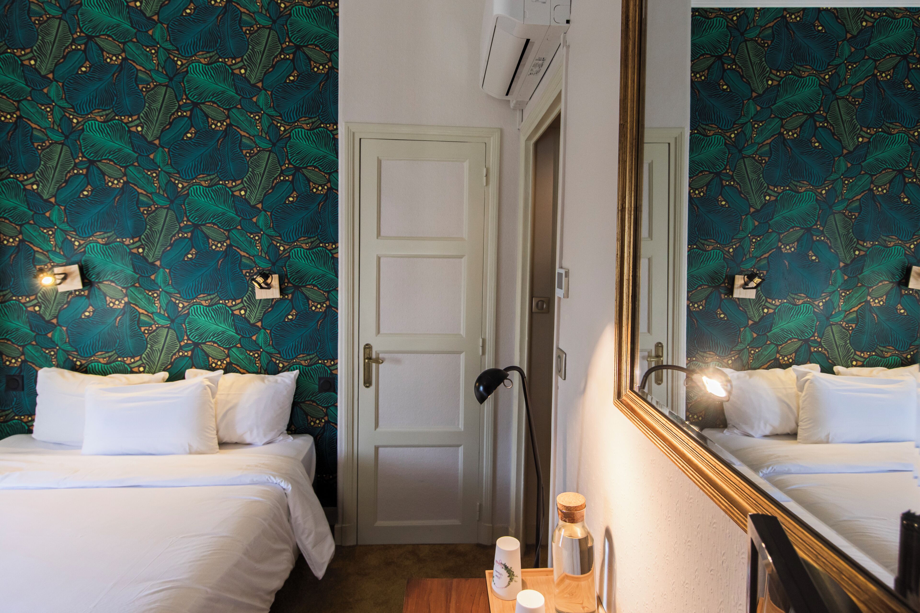 Ein Hotelzimmer mit zwei Betten, einer Tür, einem Spiegel, einer Lampe und einem Nachttisch mit einer Flasche und einem Becher.