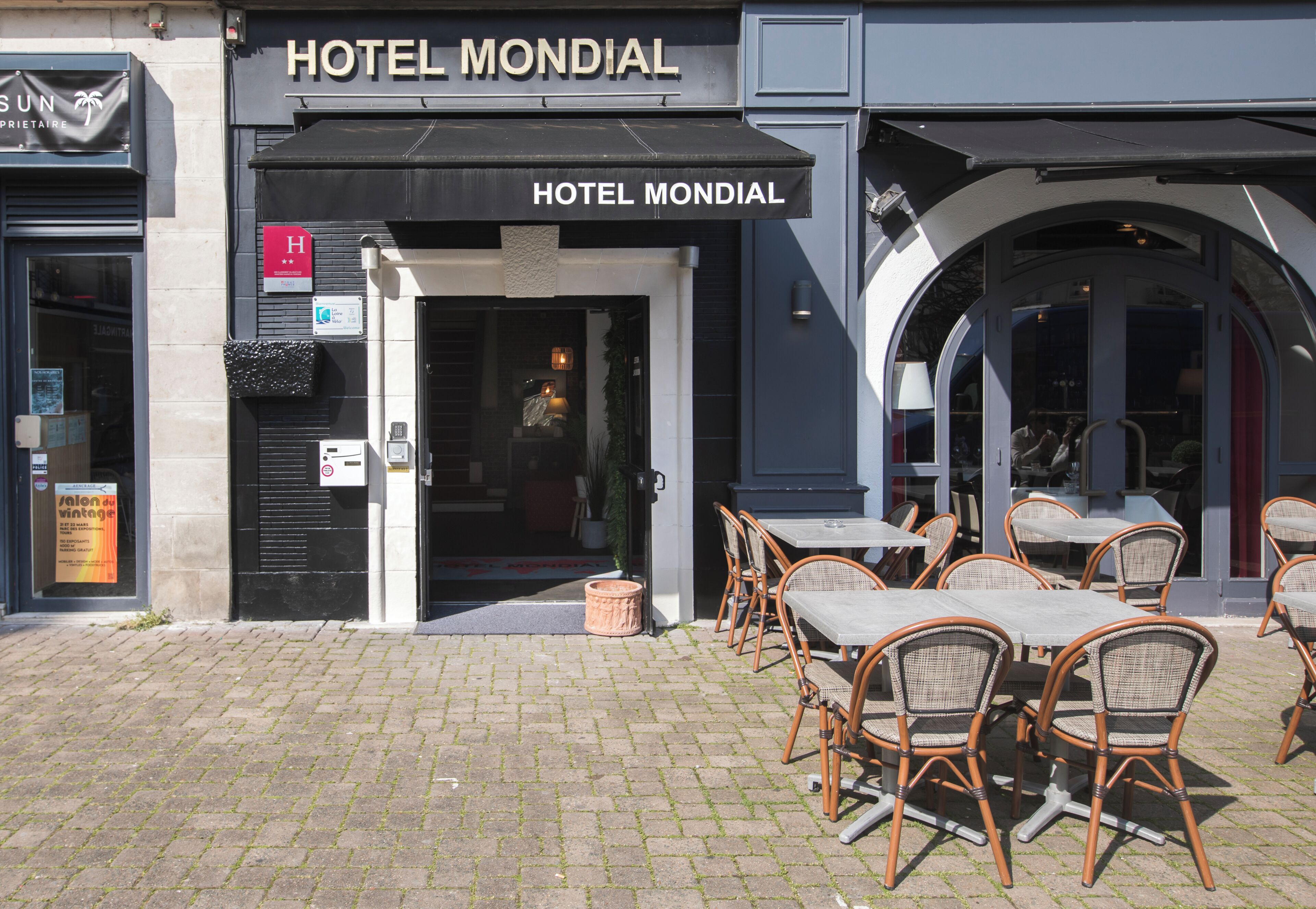 A hotel entrance with a sign reading 'HOTEL MONDIAL,' outdoor seating with wicker chairs and tables, and a cobblestone pavement.