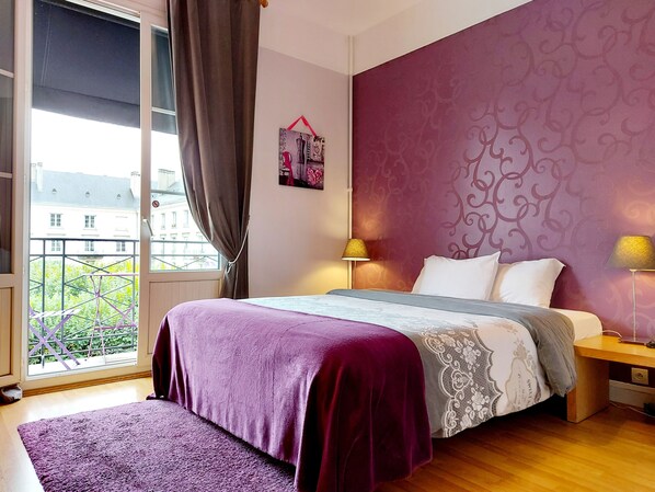 A bedroom with a bed, two bedside lamps, a window with curtains, a view of buildings, and a patterned wall. - Hotel Mondial (Tours)