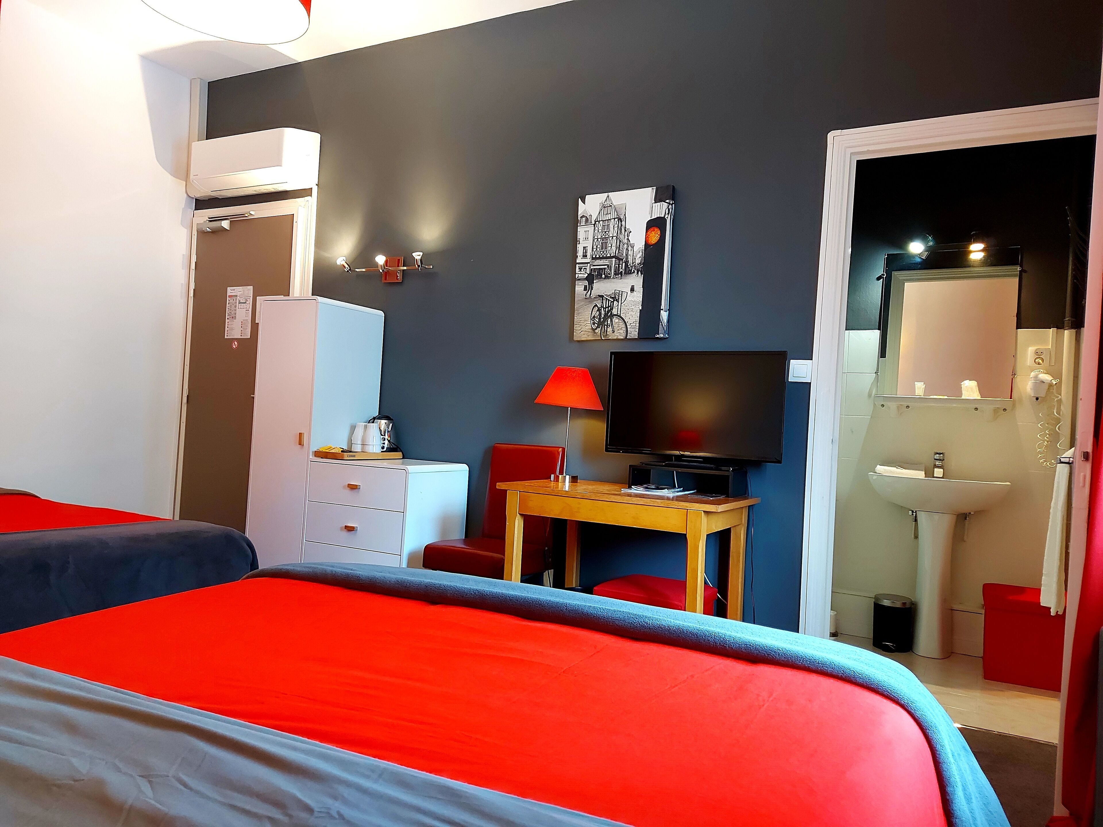 Hotellihuone, jossa on sänky, kirjoituspöytä, jossa on televisio, punainen tuoli, vaatekaappi ja kylpyhuone, joka näkyy avoimesta ovesta.