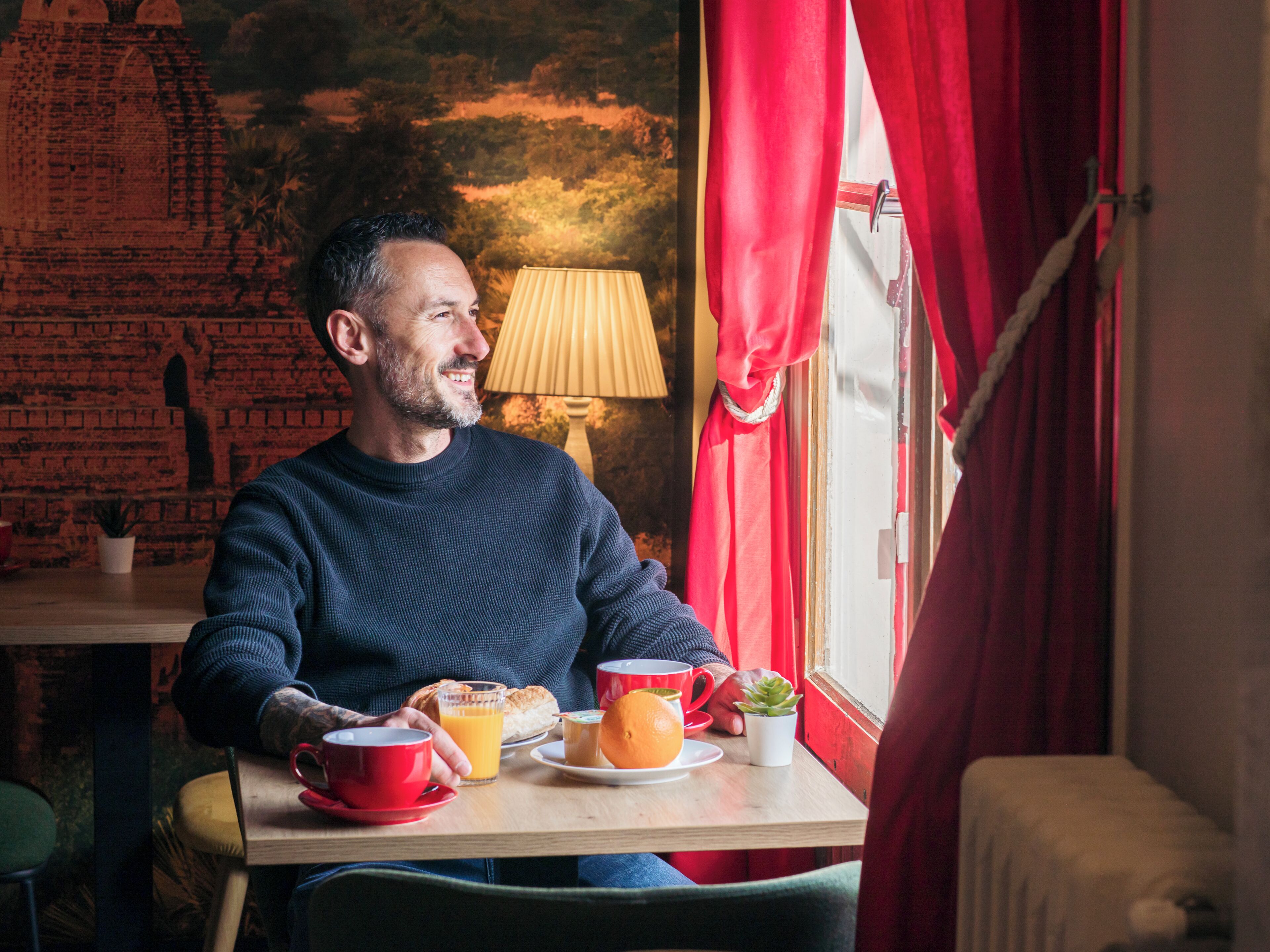 Un homme est assis à une table, devant un petit-déjeuner, une lampe et une fenêtre ornée de rideaux rouges.