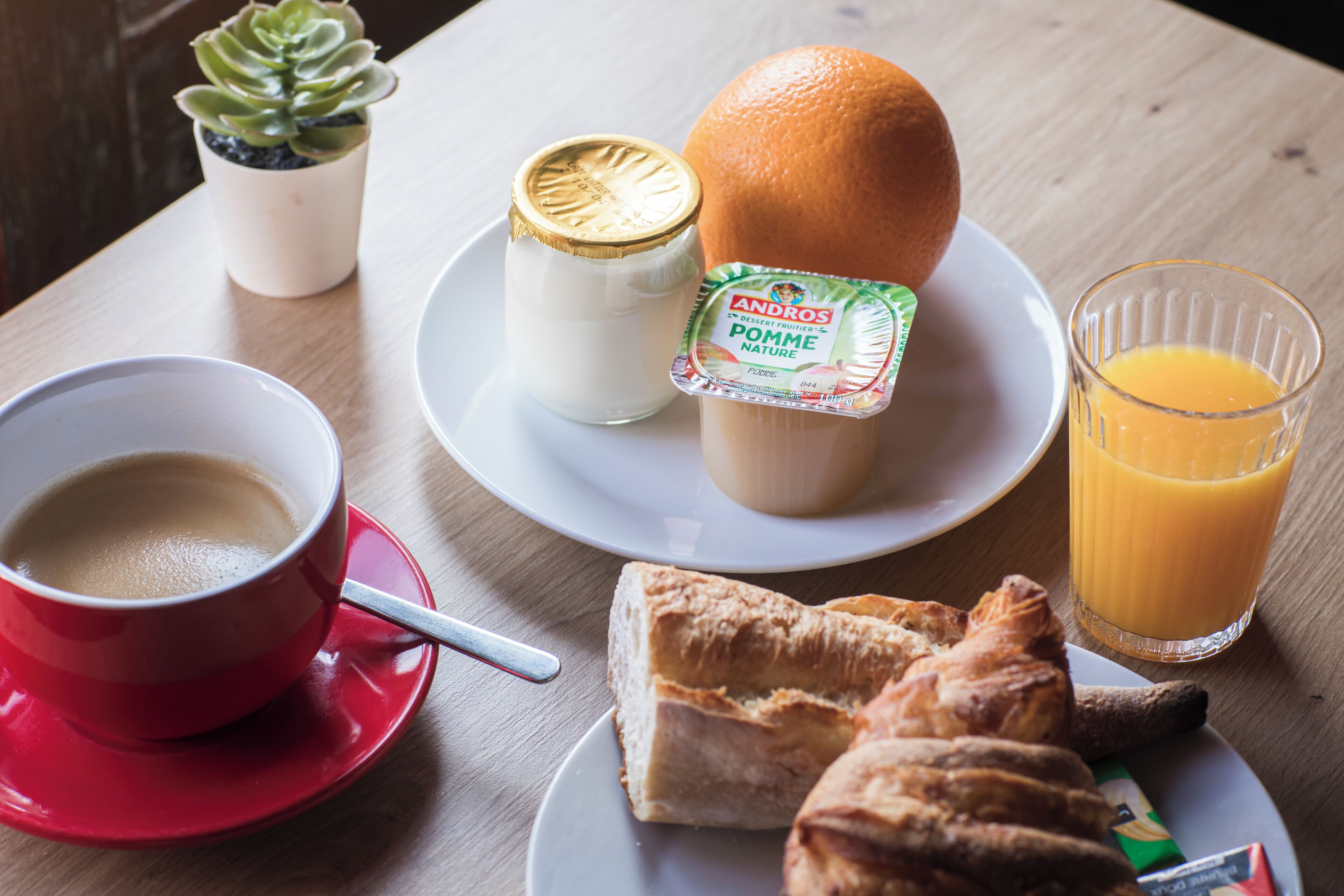 A breakfast setting with a cup of coffee, a glass of orange juice, a croissant, a small orange, a container of Andros Pomme Nature yogurt, and a potted plant.