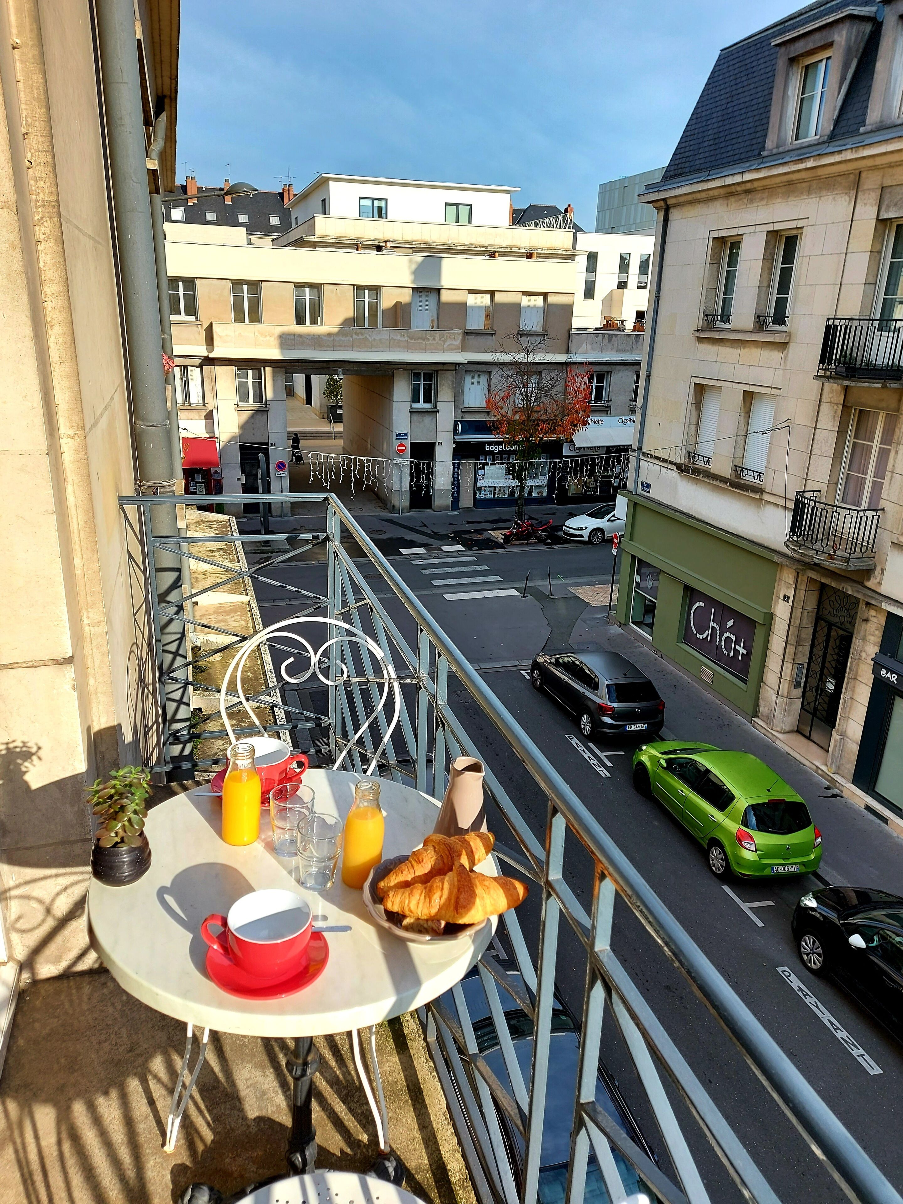 Un balcon avec une table préparée pour le petit-déjeuner, donnant sur une rue animée avec des voitures et des immeubles.