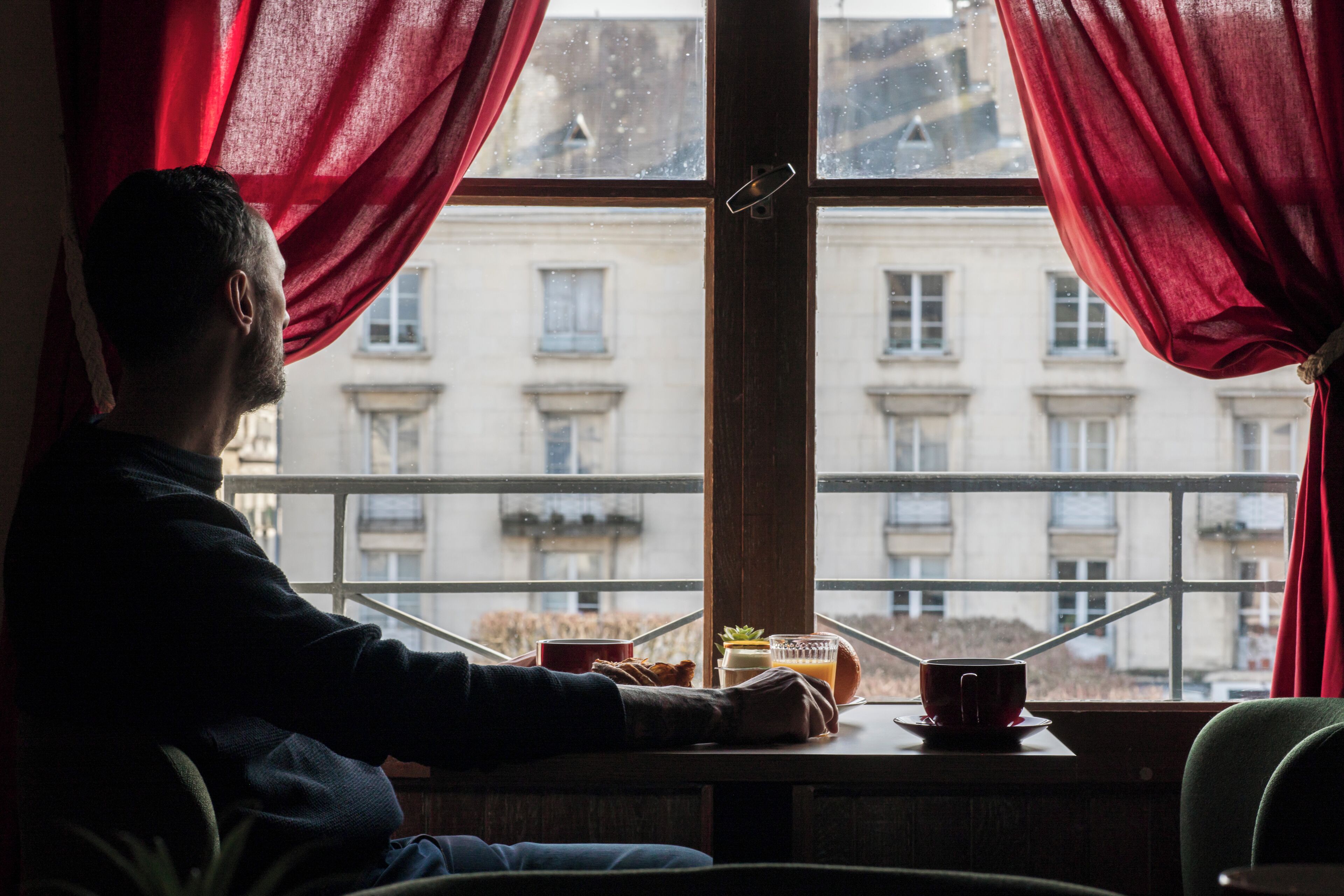 Una persona sentada junto a una ventana con cortinas rojas, una mesa con una taza, una planta pequeña y vistas de edificios desde la ventana.