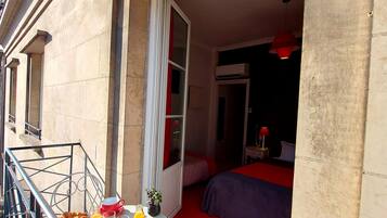 Un balcon avec une table préparée pour le petit-déjeuner, un lit avec une literie rouge et une fenêtre avec des rideaux.