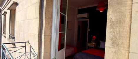 Un balcón con una mesa lista para el desayuno, una cama con ropa de cama roja y una ventana con cortinas.