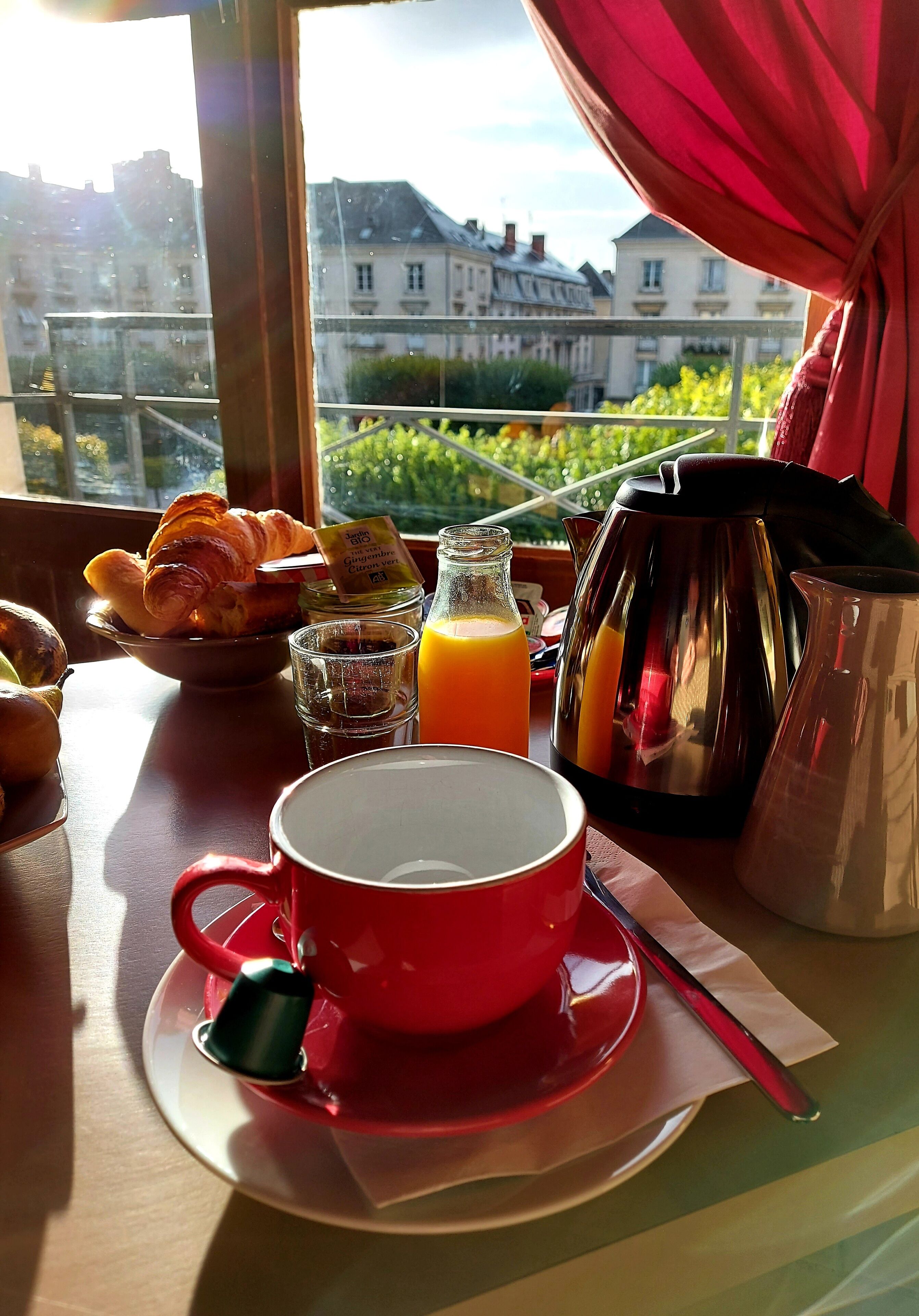 A breakfast table with croissants, a glass of orange juice, a red coffee cup, a teapot, and a pitcher.