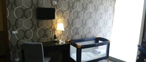 A room with a bunk bed, a desk with a lamp, a TV mounted on the wall, and a patterned wallpaper.