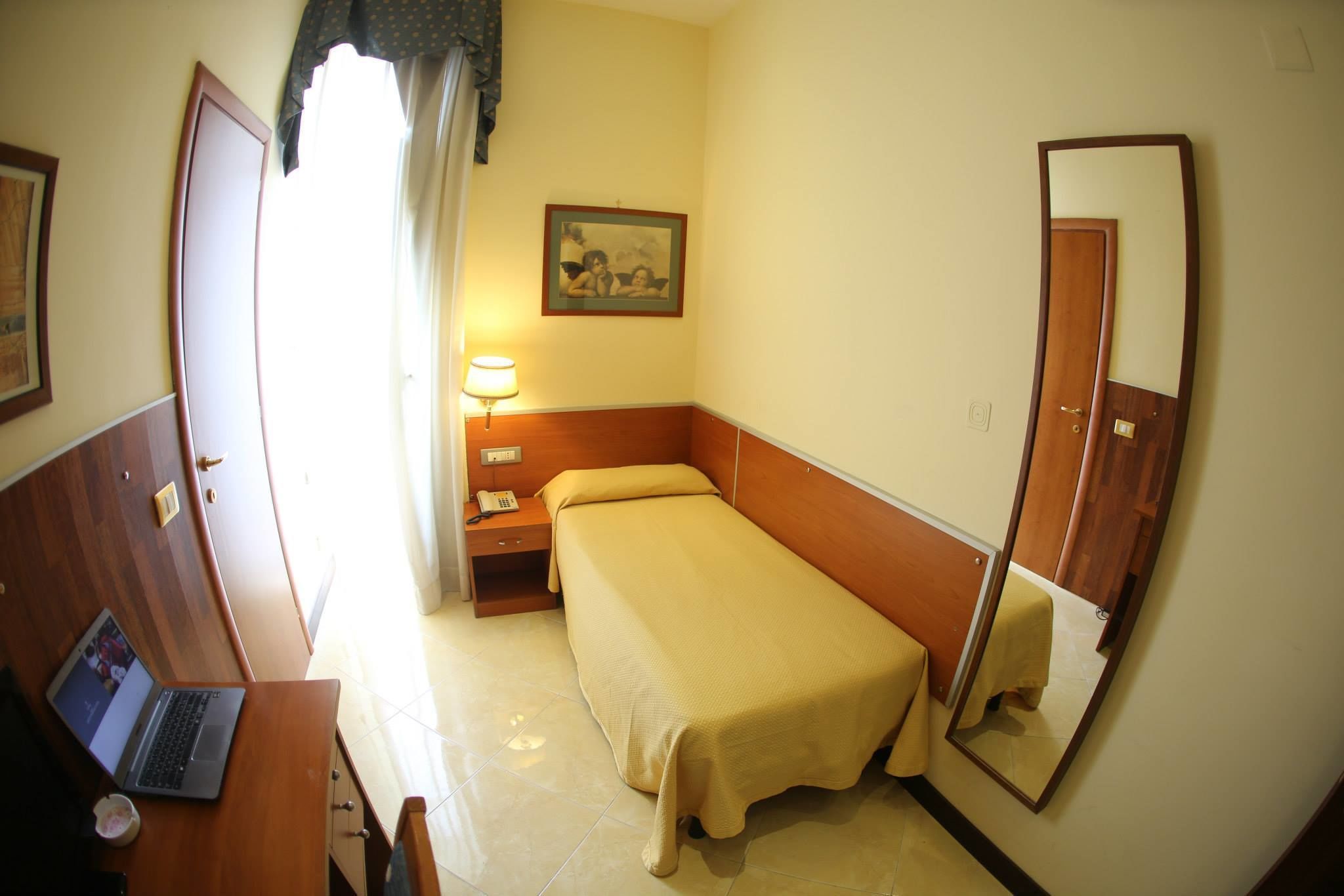 A hotel room with a bed, a desk with a laptop, a bedside table with a lamp, and a mirror on the wall.