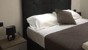 Una cama bien tendida con ropa de cama blanca y gris, un cabecero oscuro y una mesita de noche con una lámpara.